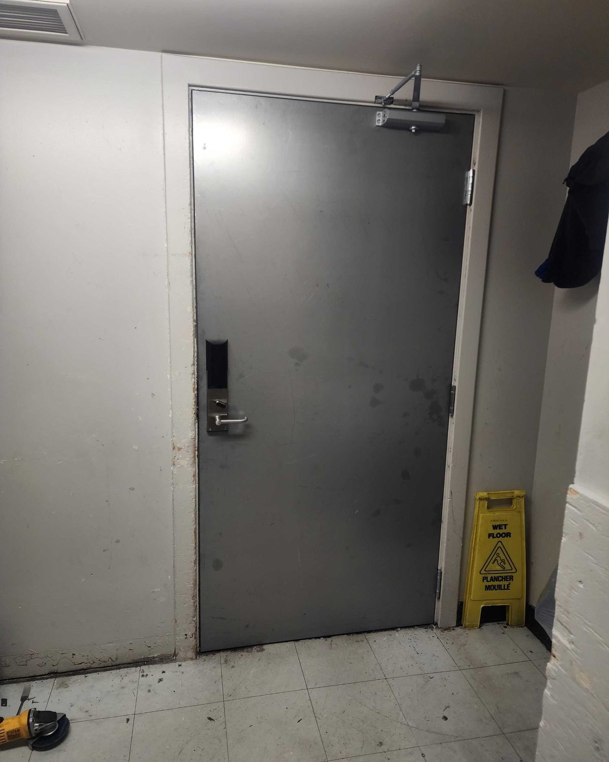 A grey steel door in a white-painted back-of-house corridor with an electronic card-reader lockset on the leaf and a surface closer at the head