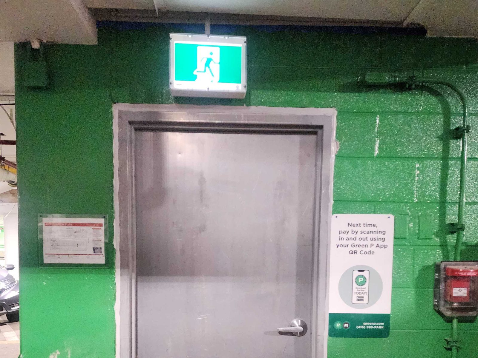 An unpainted steel door with a lever handle in a steel frame, set in a green-painted block wall of an underground parking garage under an illuminated green running-man exit sign, with an evacuation plan on one side and a fire alarm pull station on the other