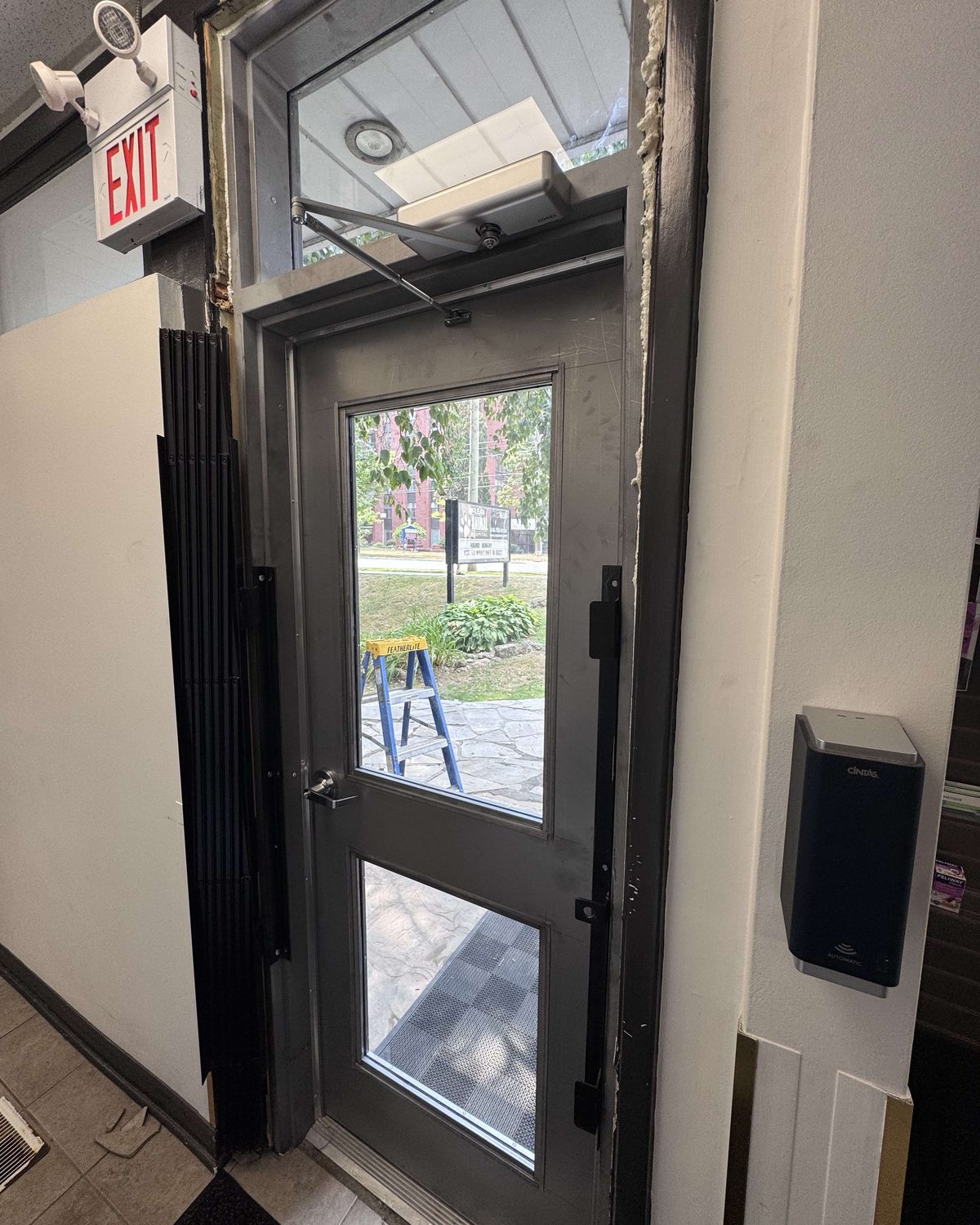 A steel door with a glass vision lite under an exit sign, a surface closer at the head and a wall-mounted card reader beside the frame