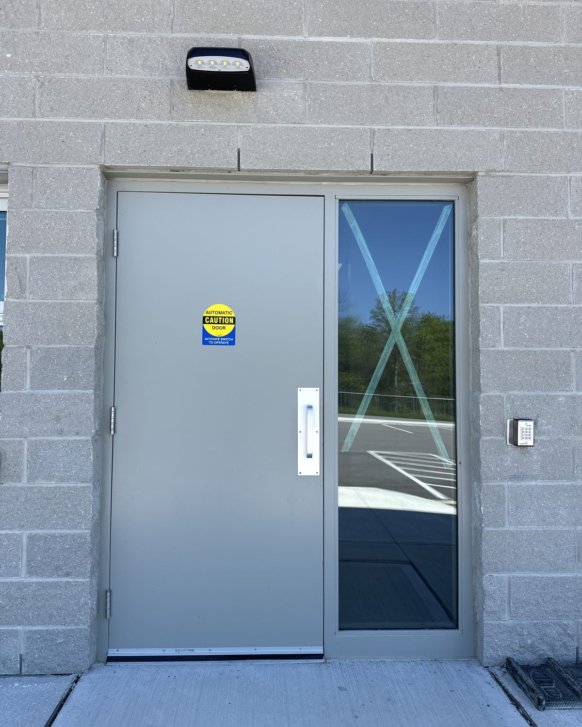 A grey steel exterior door with an automatic-door decal, a long pull handle and a tall glass sidelite, set in a split-face block wall with a keypad on the right