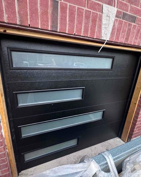 A new black sectional garage door with long horizontal frosted-glass inserts, installed in a red brick opening with the wood buck still unfinished around it