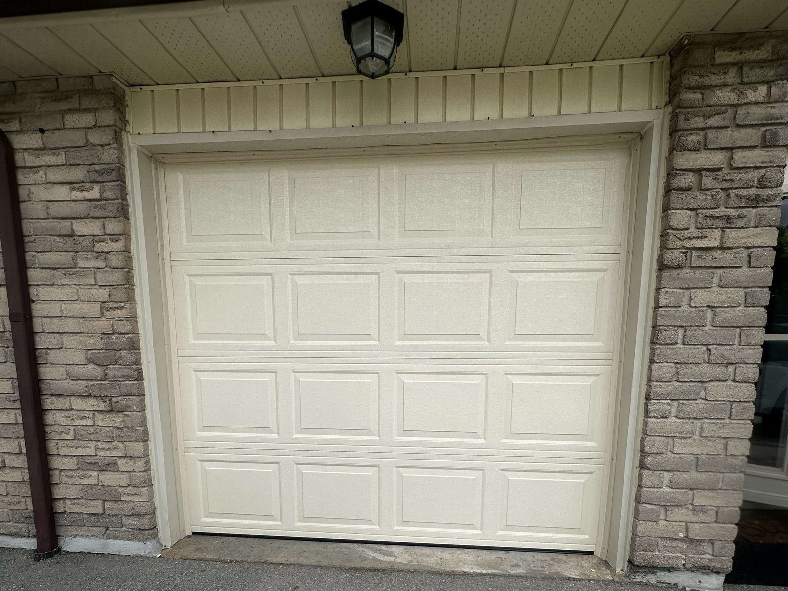 A single cream raised-panel sectional garage door, four rows of short panels, set into a grey brick opening under a white soffit with a black lantern light above it