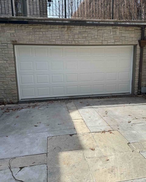 A white double-width sectional garage door with rows of rectangular raised panels, set into a buff stone wall above a flagstone courtyard