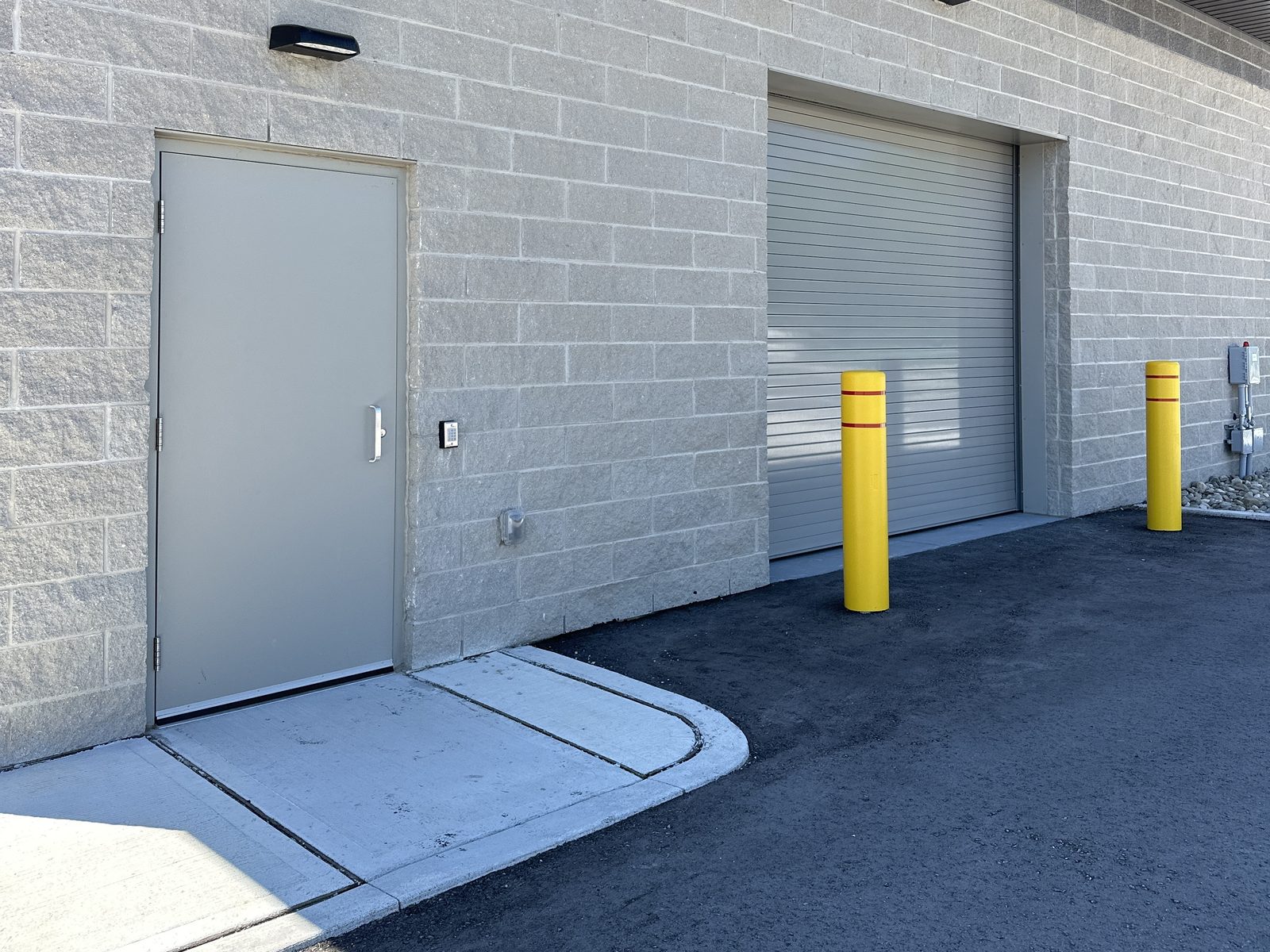 A grey steel service door with a pull handle and a keypad beside it, set in a split-face block wall of a new industrial building, beside a closed rolling steel door guarded by two yellow bollards
