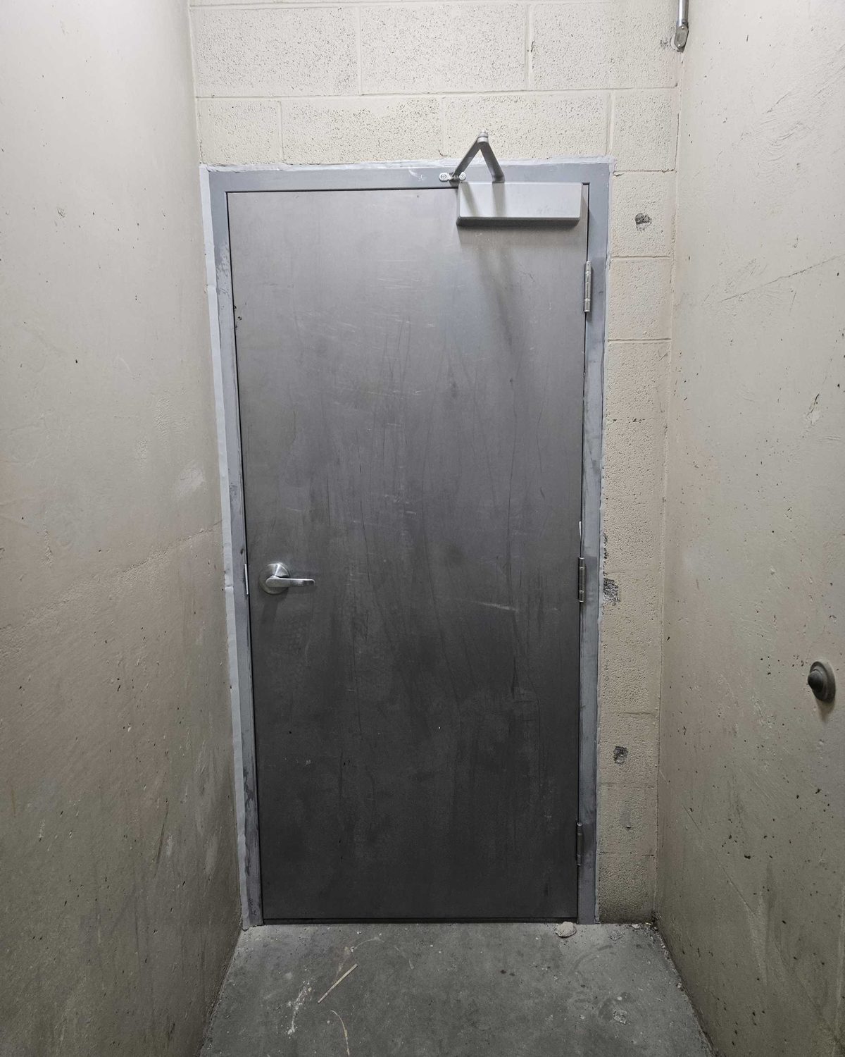 A dark grey steel door with a surface-mounted closer and a lever handle, hung in a steel frame at the end of a narrow concrete alcove