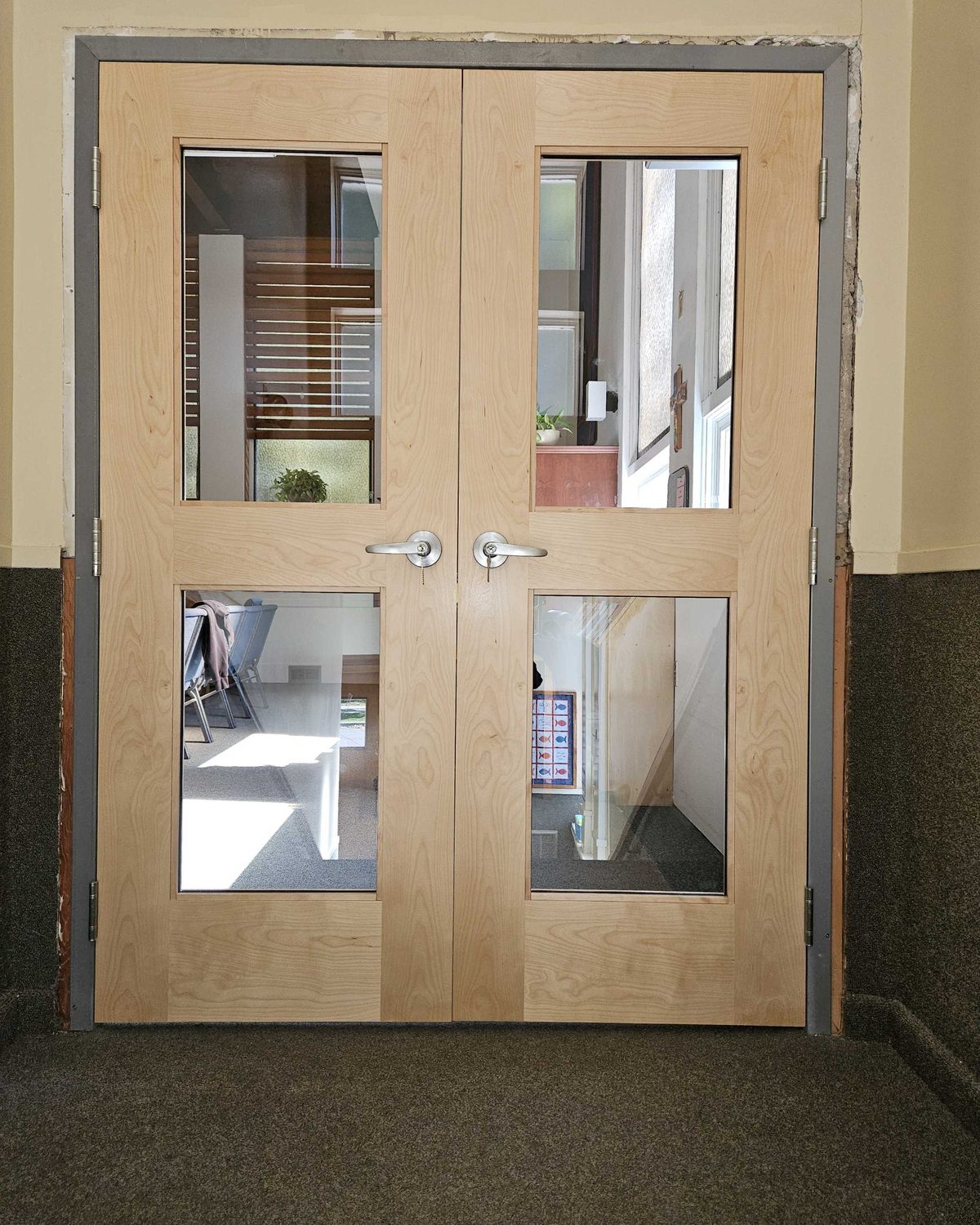 A pair of light-coloured wood stile-and-rail doors with four large clear glass panels and lever handles, hung in a grey steel frame in a church hallway