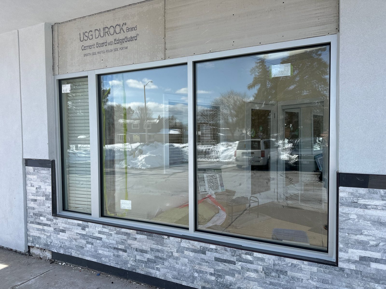 A new three-lite aluminum storefront window at a Mississauga plaza in winter, the glass still carrying its paper labels, above a grey stacked-stone base, with snow banks and parked cars reflected in the lites