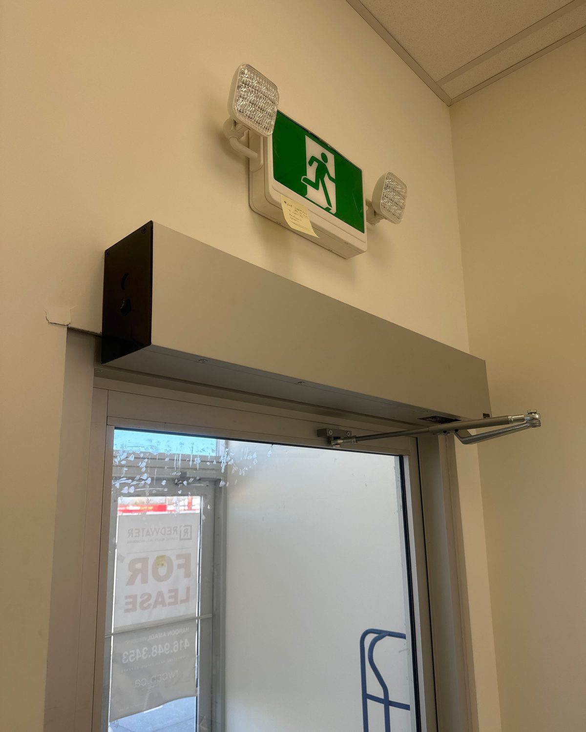 A surface-mounted swing door operator in a silver header box above a glazed aluminum door, its drive arm connected to the top of the leaf, beneath an exit sign with emergency lights