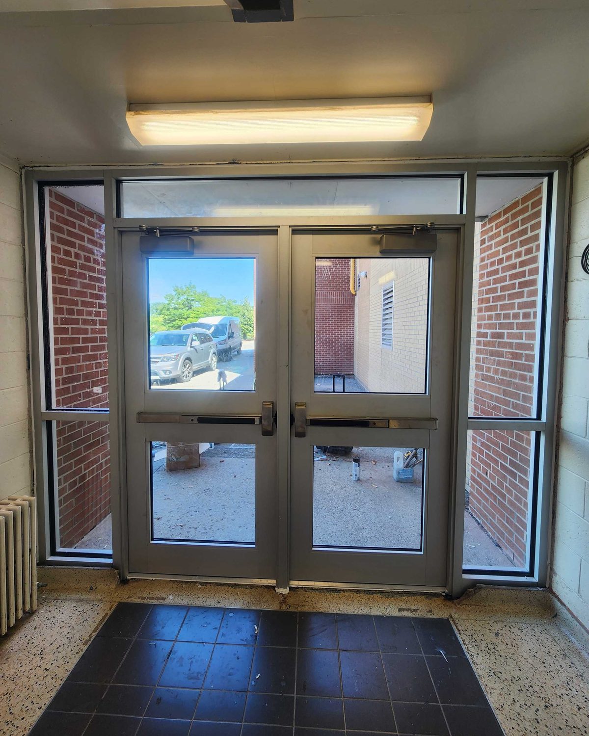 A pair of wider-stile aluminum doors with a mid rail and exit devices, overhead closers on the top rail, glazed sidelites and a transom, seen from inside a brick vestibule