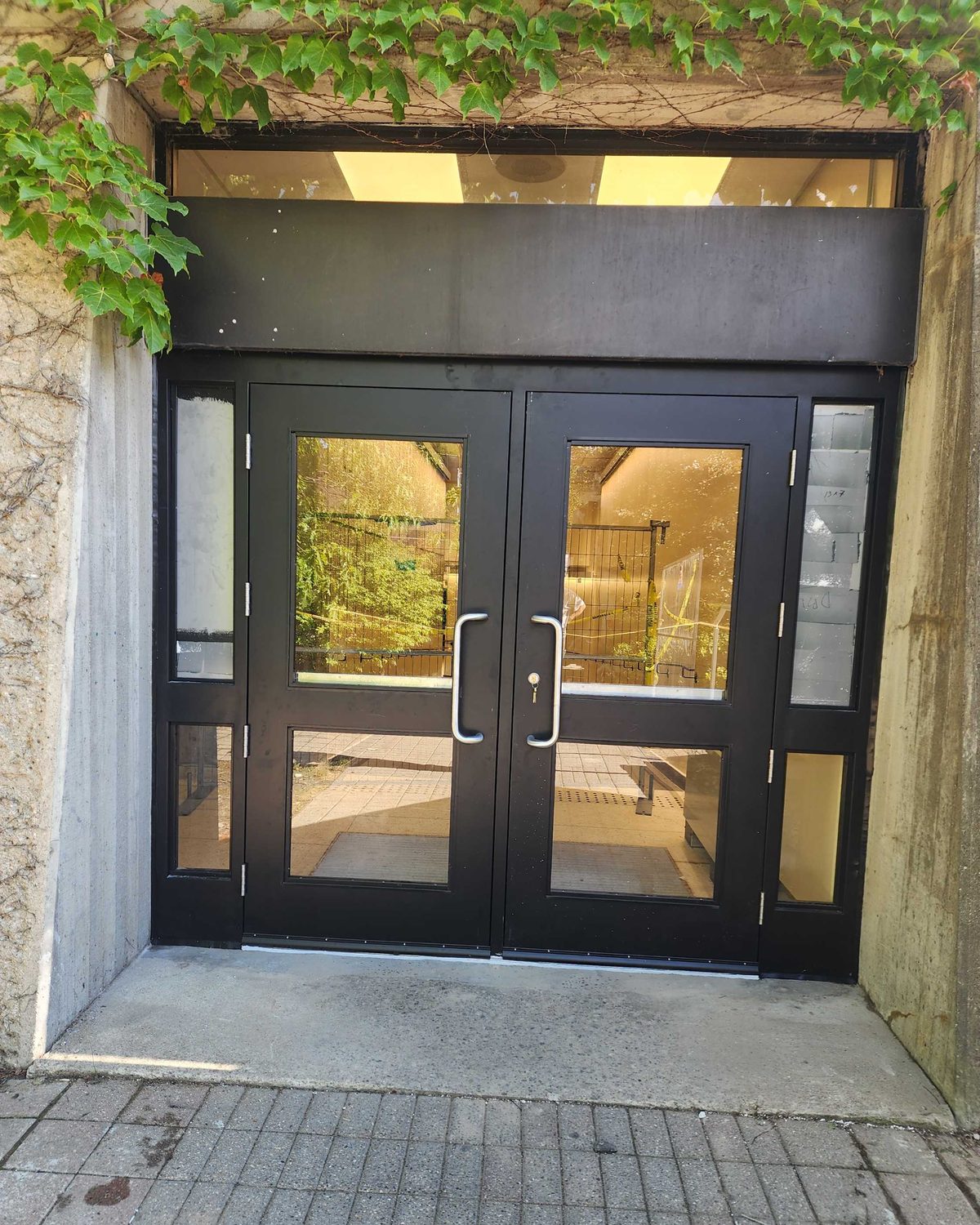 Black-anodized wide-stile aluminum entrance door pair with narrow sidelites and a transom in a concrete surround, installed by Canuck Door Systems