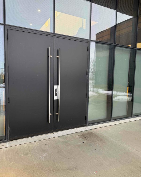 A pair of dark aluminum entrance doors with full-height pull handles and a centre cylinder, set into a glazed curtain wall, supplied and installed by Canuck Door Systems