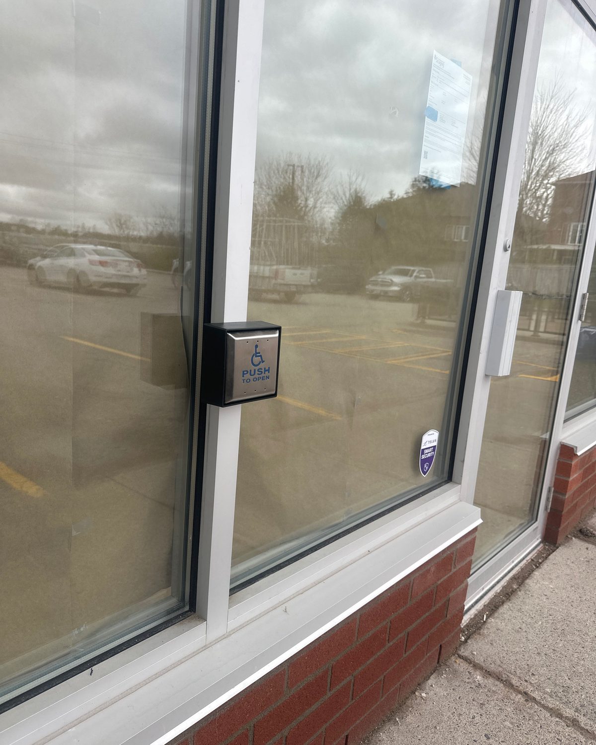 A stainless steel wall-mounted door actuator plate beside a glazed commercial entrance, installed by Canuck Door Systems