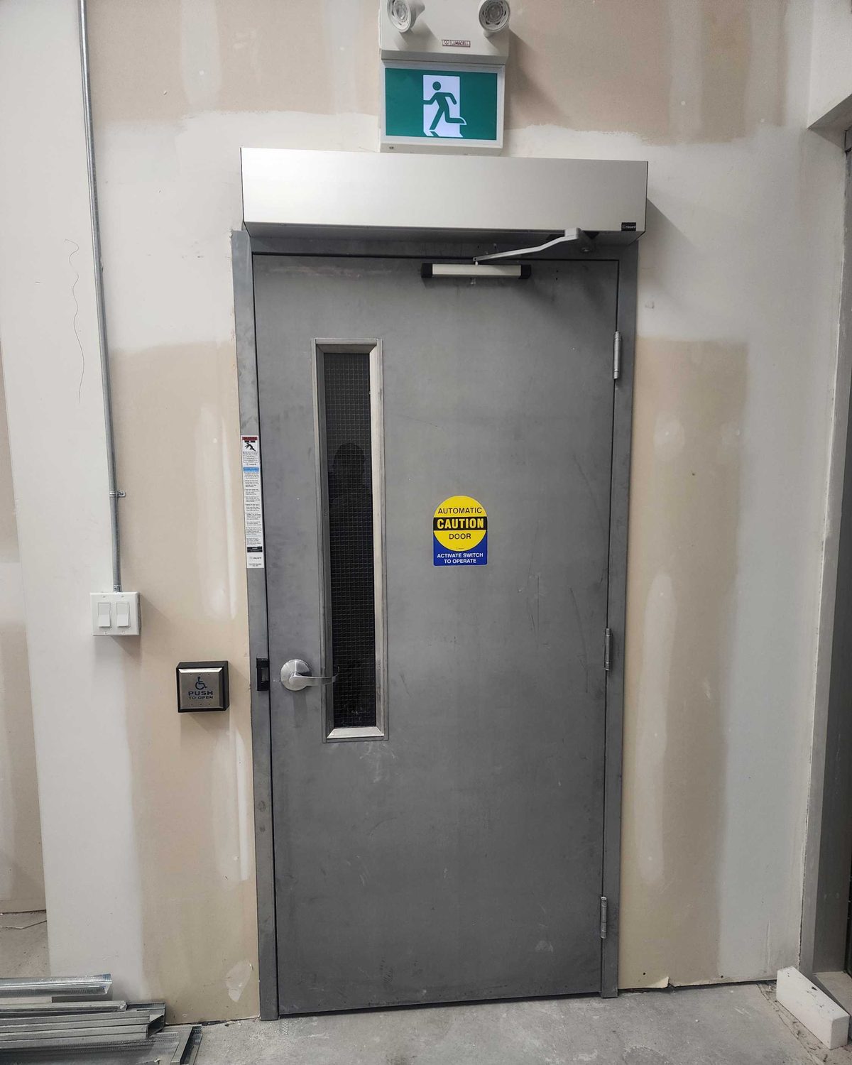 A steel door with a vision lite and a surface-mounted automatic operator above it in a commercial corridor, installed by Canuck Door Systems