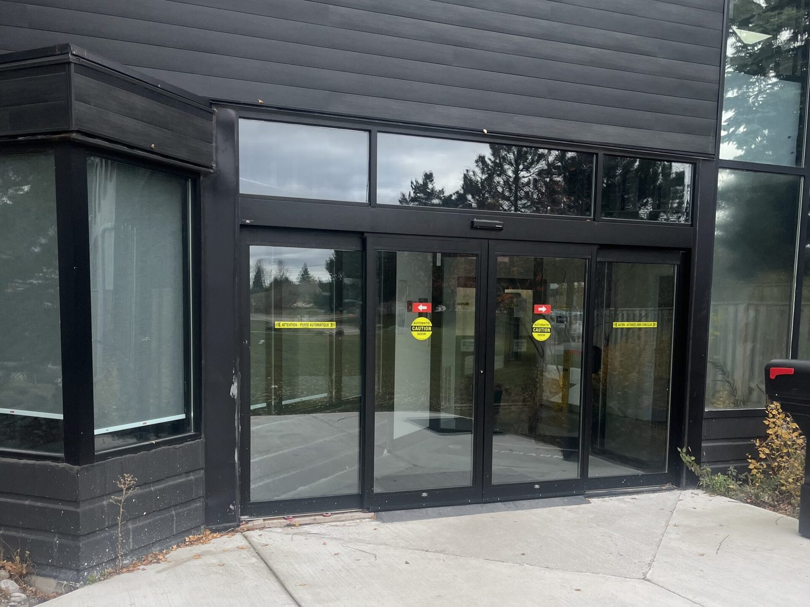 A bi-parting automatic sliding entrance in black aluminum framing with glazed sidelites and a transom above, carrying bilingual automatic-door caution decals, installed by Canuck Door Systems
