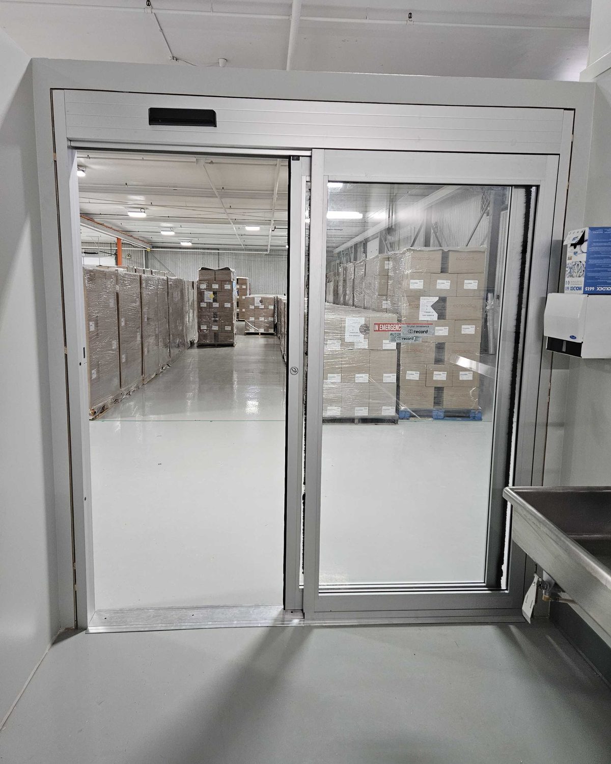 A white-framed automatic sliding door between a cleanroom and a warehouse in a food plant, with a Record automatic door decal on the glass, installed by Canuck Door Systems