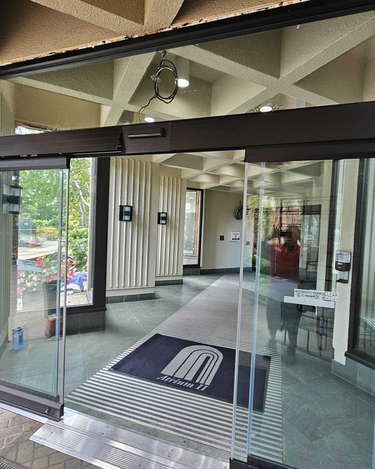 A bi-parting automatic sliding entrance seen from inside a building lobby, with an entrance mat and glazed sidelites, installed by Canuck Door Systems