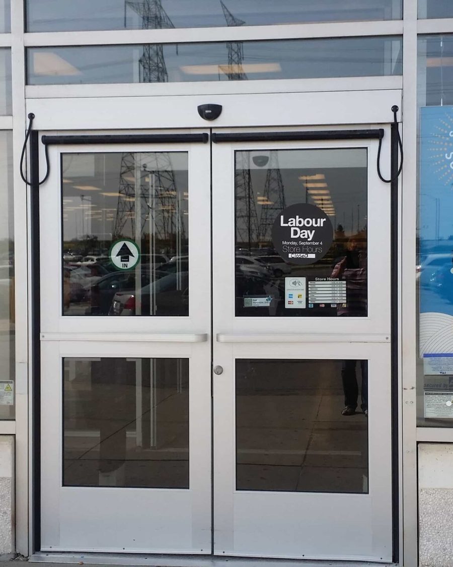 A bi-parting automatic sliding entrance in aluminum framing with guide rails and a header-mounted sensor at a retail storefront, installed by Canuck Door Systems