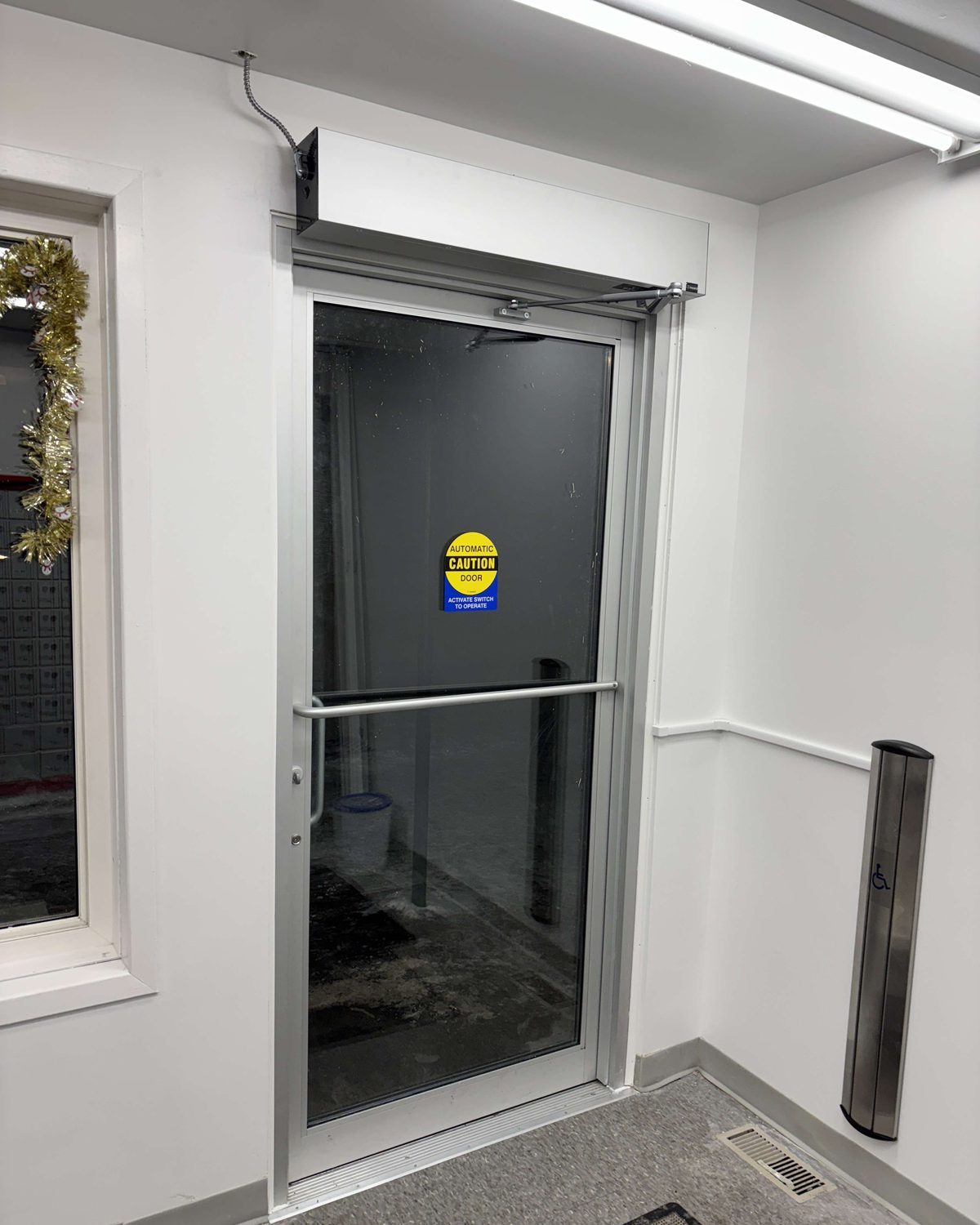 A surface-mounted door operator above a glazed interior door with a horizontal push bar and a floor-mounted actuator post, installed by Canuck Door Systems