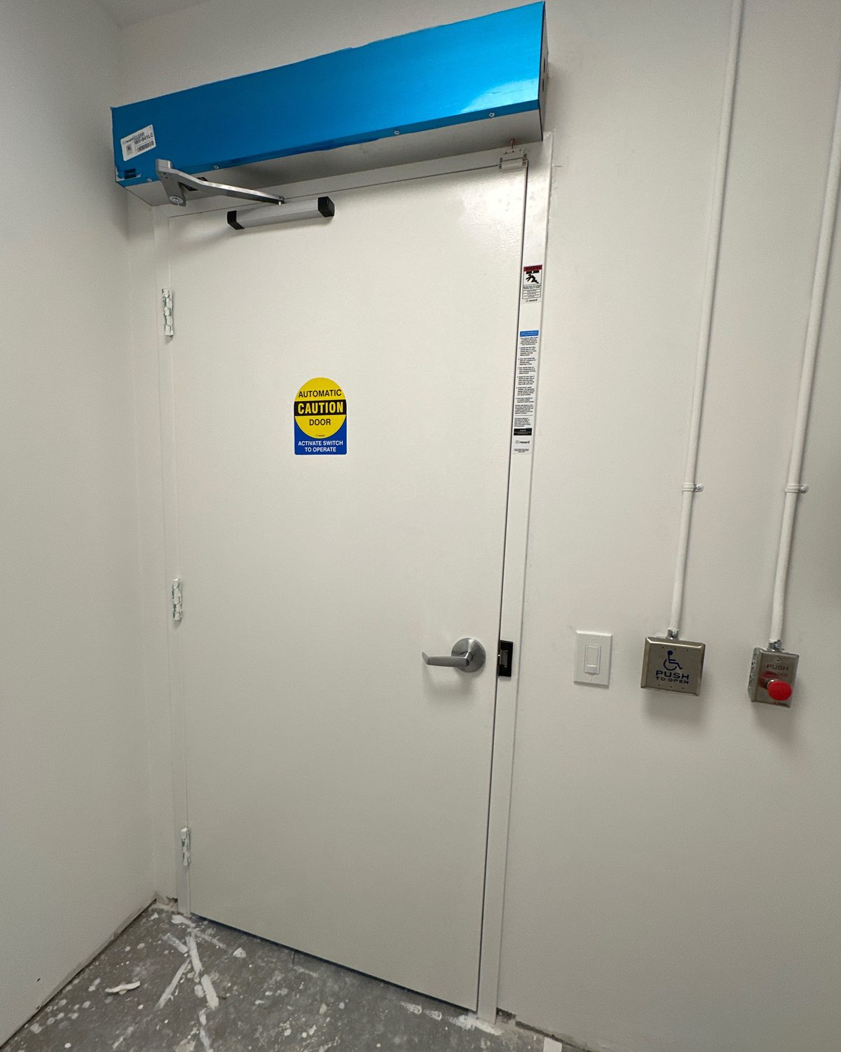 A low-energy door operator in a blue header cover above a steel door, with wall-mounted actuator switches beside it, installed by Canuck Door Systems