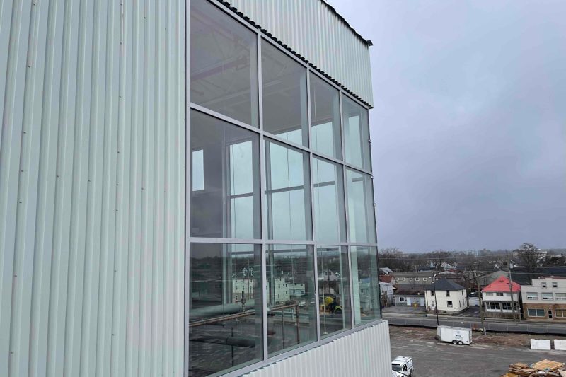 A two-storey glazed corner in white aluminum framing, carried past the floor line and set into the metal-clad elevation of an industrial building, installed by Canuck Door Systems
