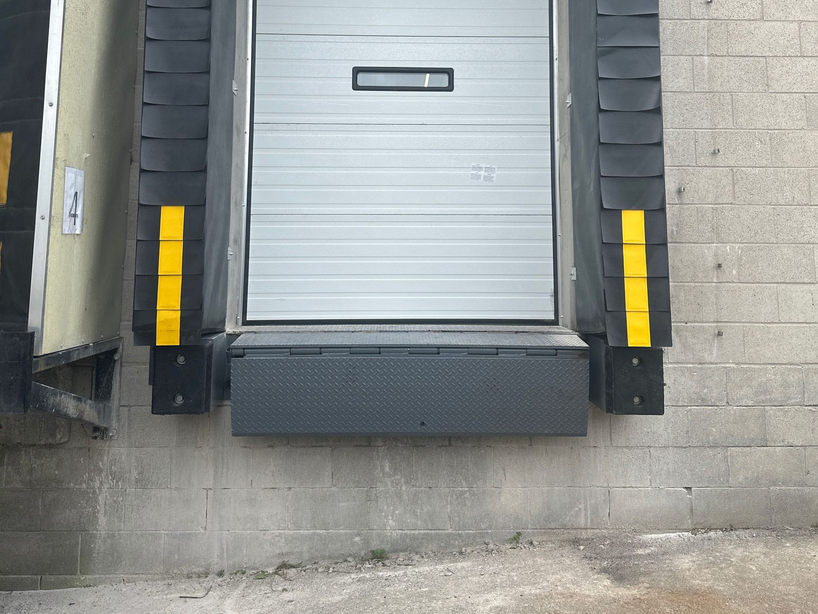 A diamond-plate edge-of-dock leveler mounted at a dock position between black laminated bumpers, under a white sectional door, installed by Canuck Door Systems