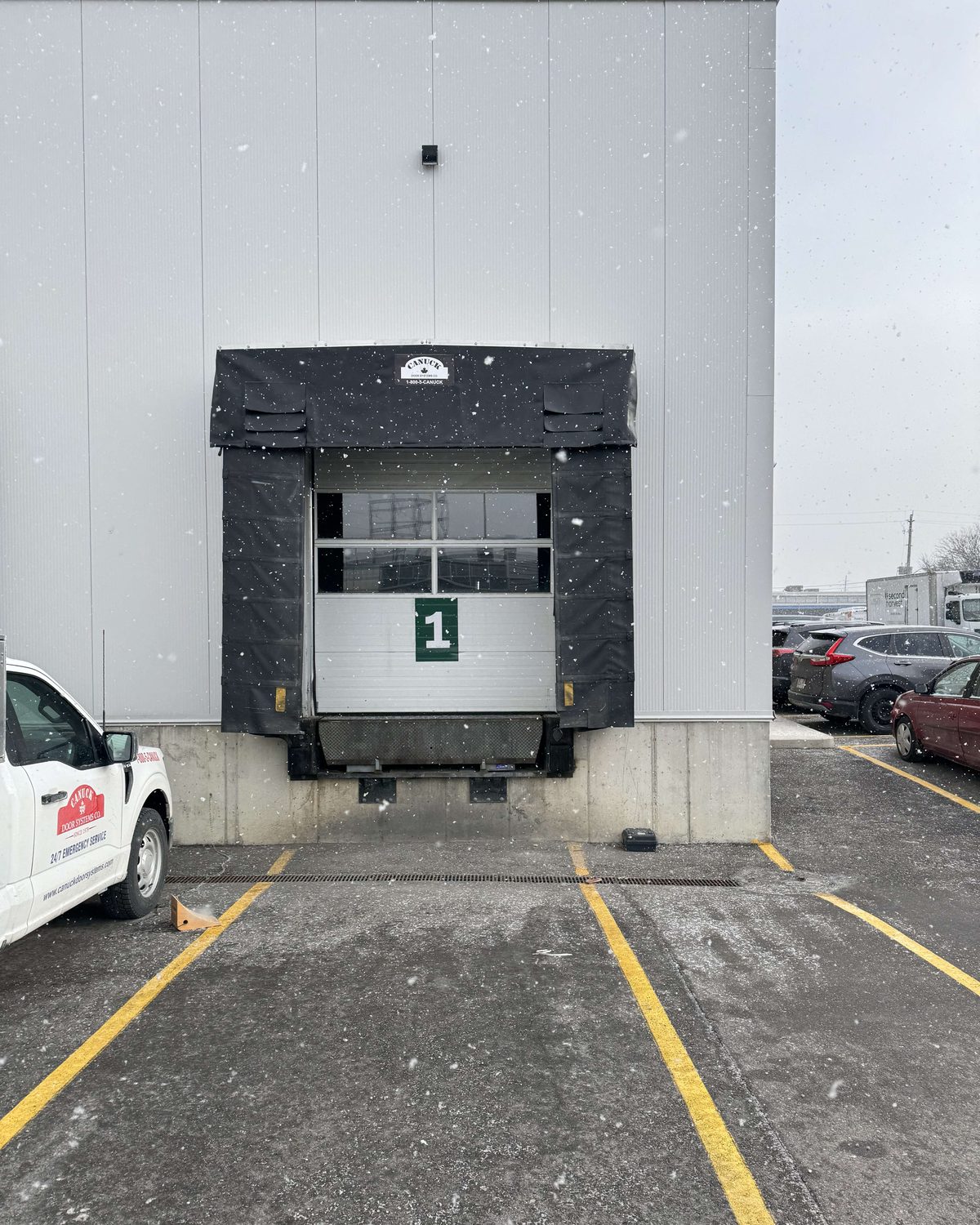 A black compression dock seal with head and side pads around a white sectional dock door in falling snow, installed by Canuck Door Systems