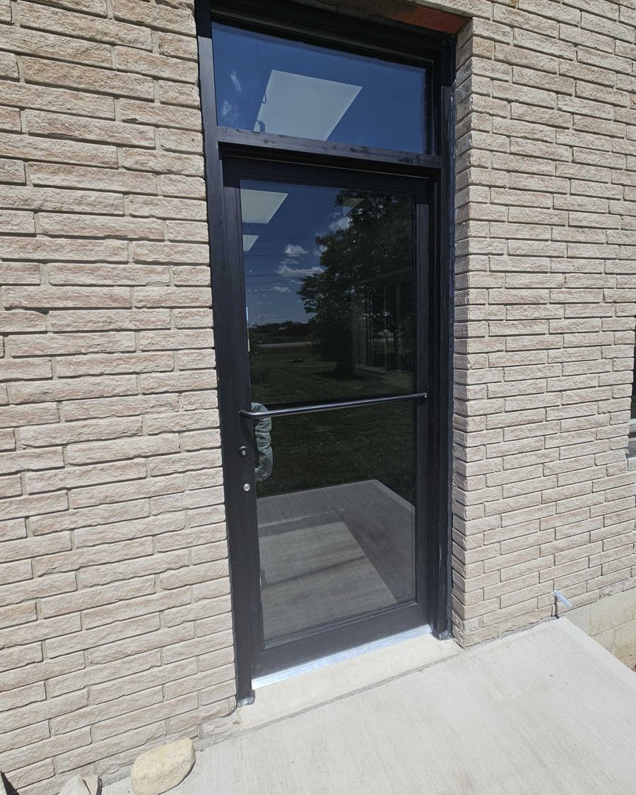 A black aluminum and glass entrance door with a push bar, a cylinder lock and a transom above, set in a brick wall, installed by Canuck Door Systems