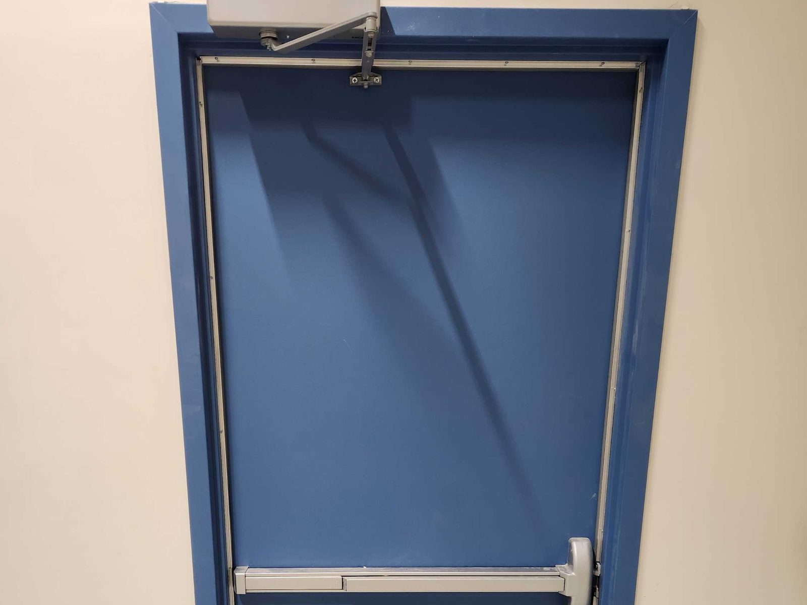 A blue steel door fitted with a horizontal exit device and a surface-mounted door closer in a commercial building, installed by Canuck Door Systems