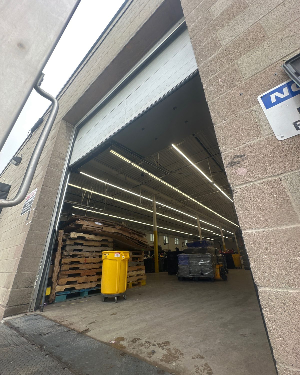 An open overhead door at a commercial warehouse bay with stacked pallets and lit racking inside, serviced by Canuck Door Systems