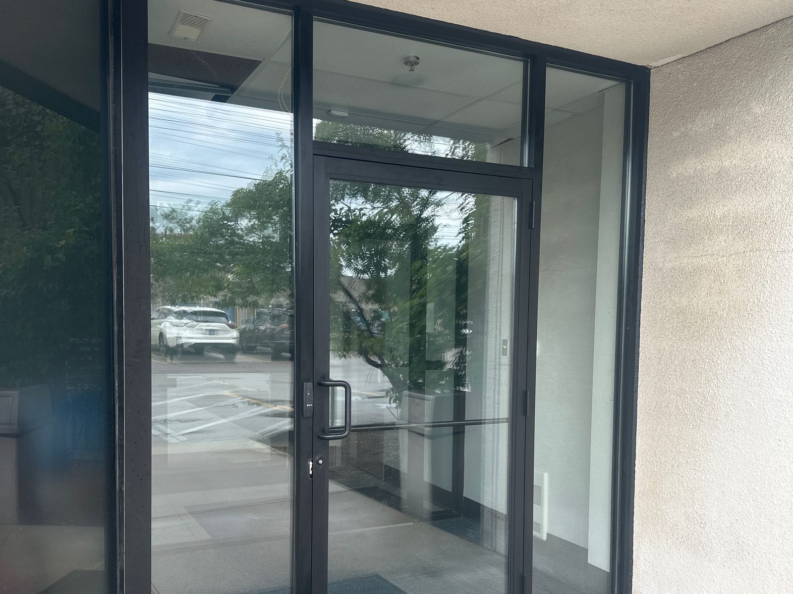 A black aluminum-framed commercial entrance door with glazed sidelites and a transom, set under a soffit and facing a parking lot, serviced by Canuck Door Systems