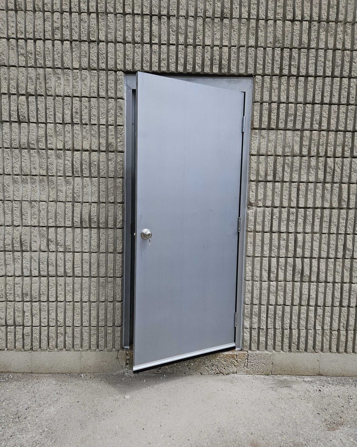 A single grey hollow metal door standing ajar in a split-face block wall at the back of a commercial building, serviced by Canuck Door Systems