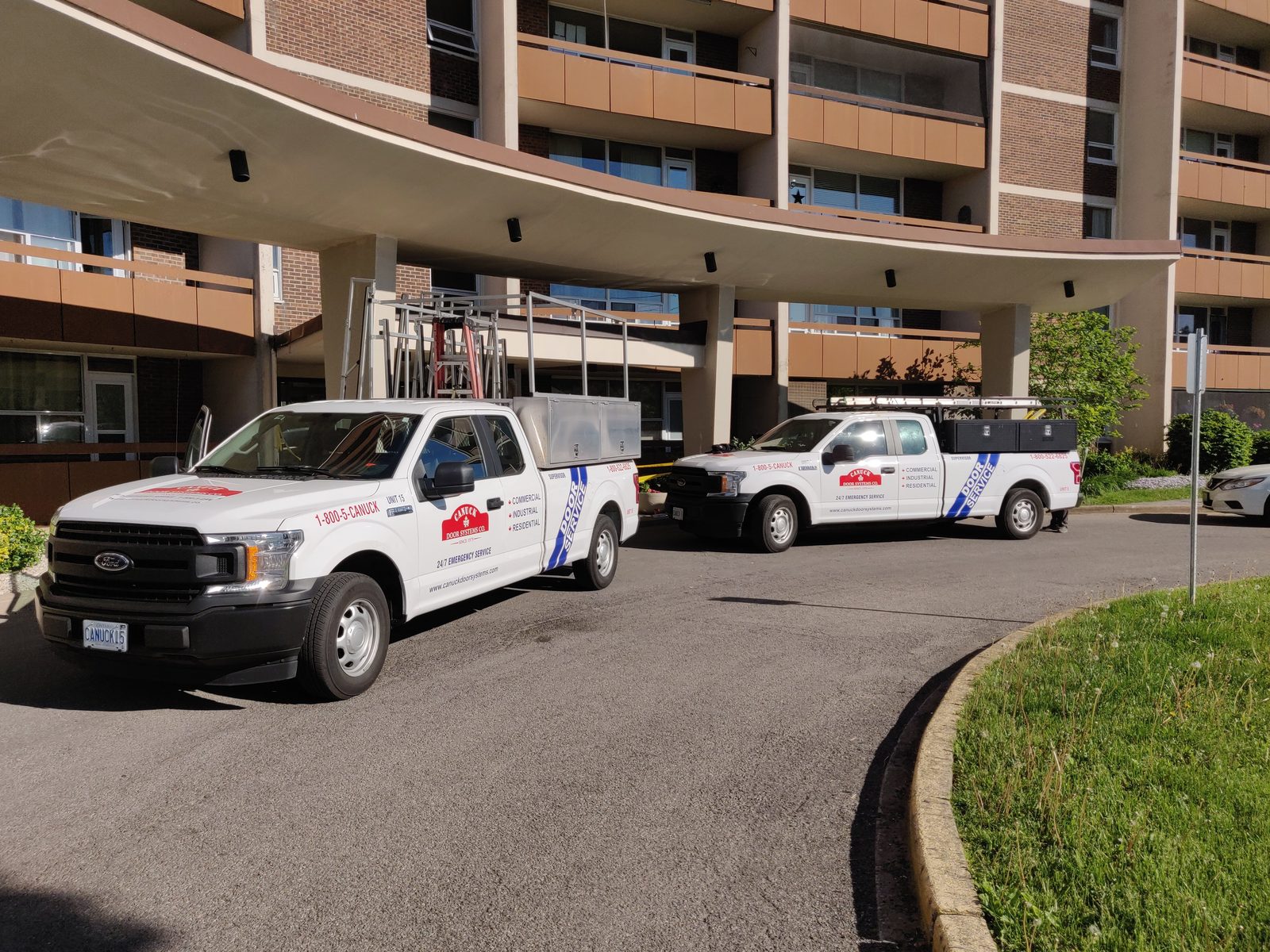 Two Canuck Door Systems service trucks parked under the canopy at an apartment building entrance, signwritten commercial, industrial, residential and 24/7 emergency service with the 1-800-5-CANUCK number