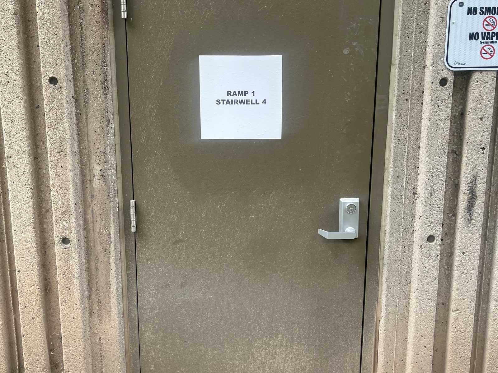 A numbered stairwell fire door with posted notices and an exit device in a concrete parking structure, inspected by Canuck Door Systems