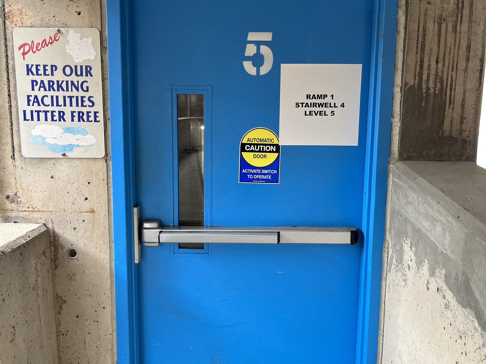 A blue stairwell door with a narrow wired-glass vision lite, exit device and level signage in a concrete parking structure, installed by Canuck Door Systems