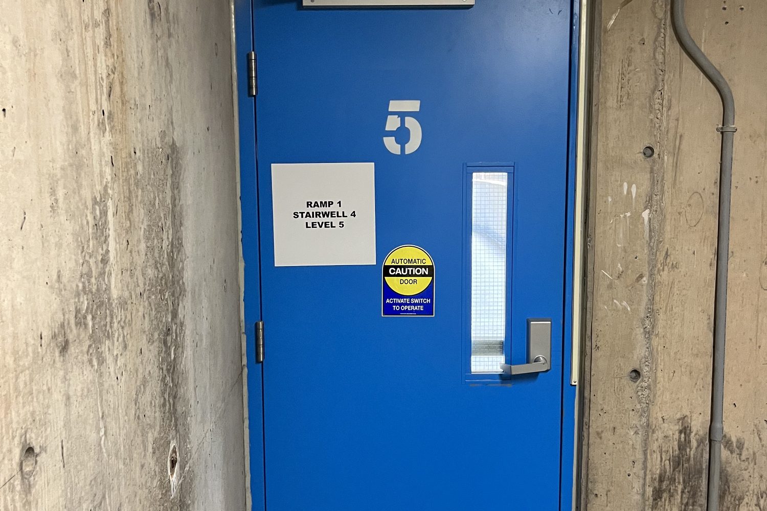 A blue-painted steel stairwell door with an exit device and level number in a concrete parking structure, installed by Canuck Door Systems