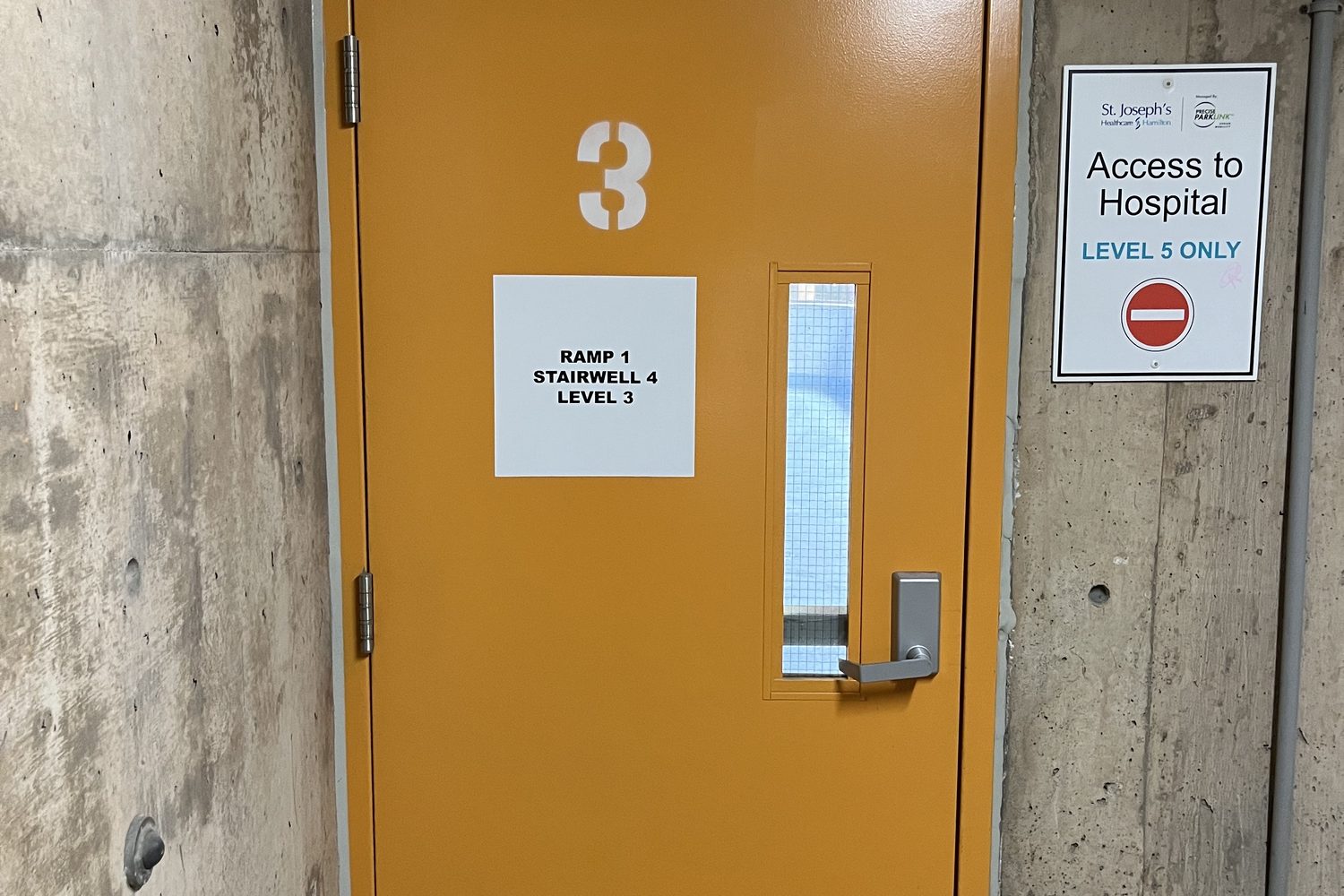 An orange-painted steel stairwell door with an exit device and hospital access signage, installed by Canuck Door Systems