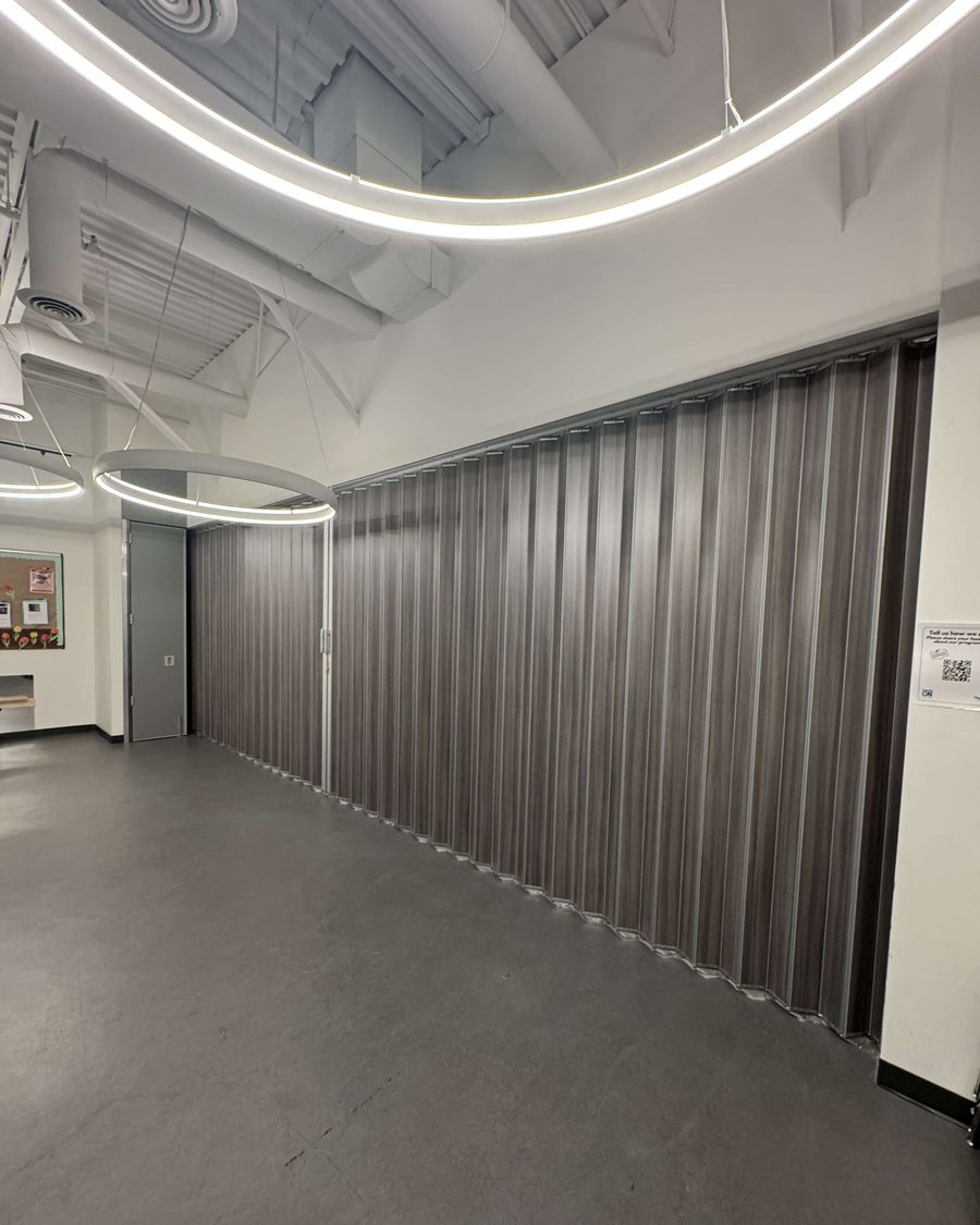 A dark grey folding accordion partition closed across a wide corridor in a community centre with curved ceiling lighting, installed by Canuck Door Systems