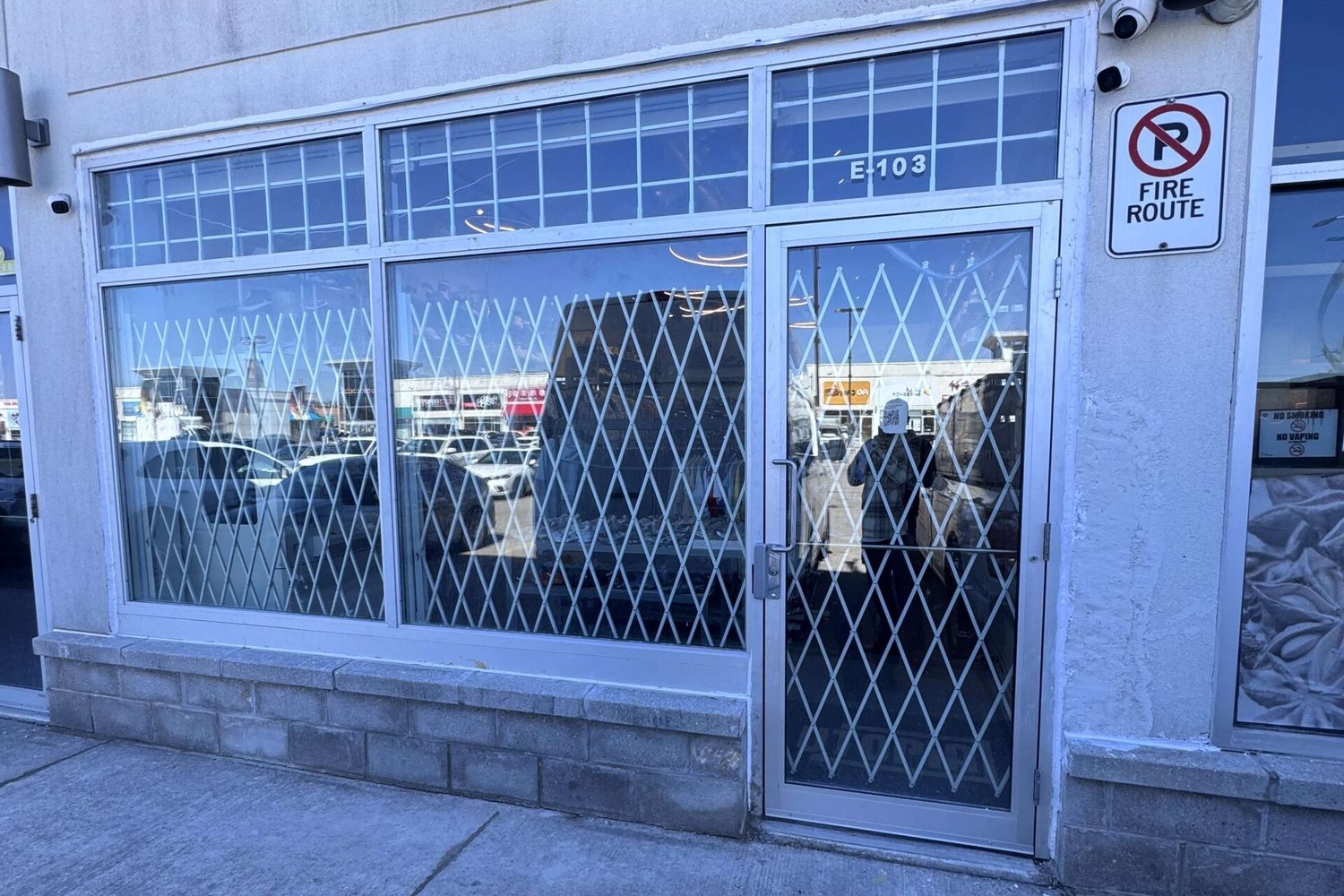 A diamond-pattern folding security gate closed behind the glazing and entrance door of a plaza storefront, installed by Canuck Door Systems