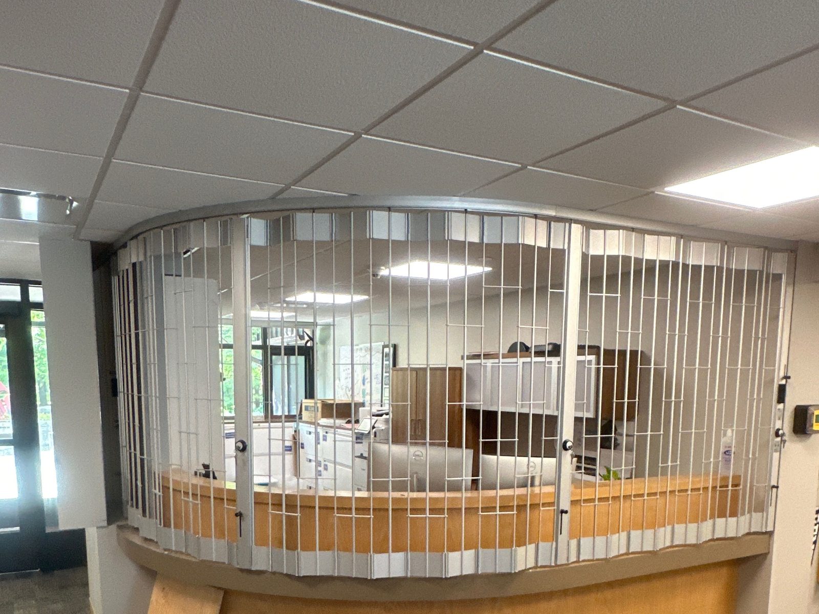 A folding security grille closed across a curved reception counter in a school, its vertical picket sections following the bend, installed by Canuck Door Systems
