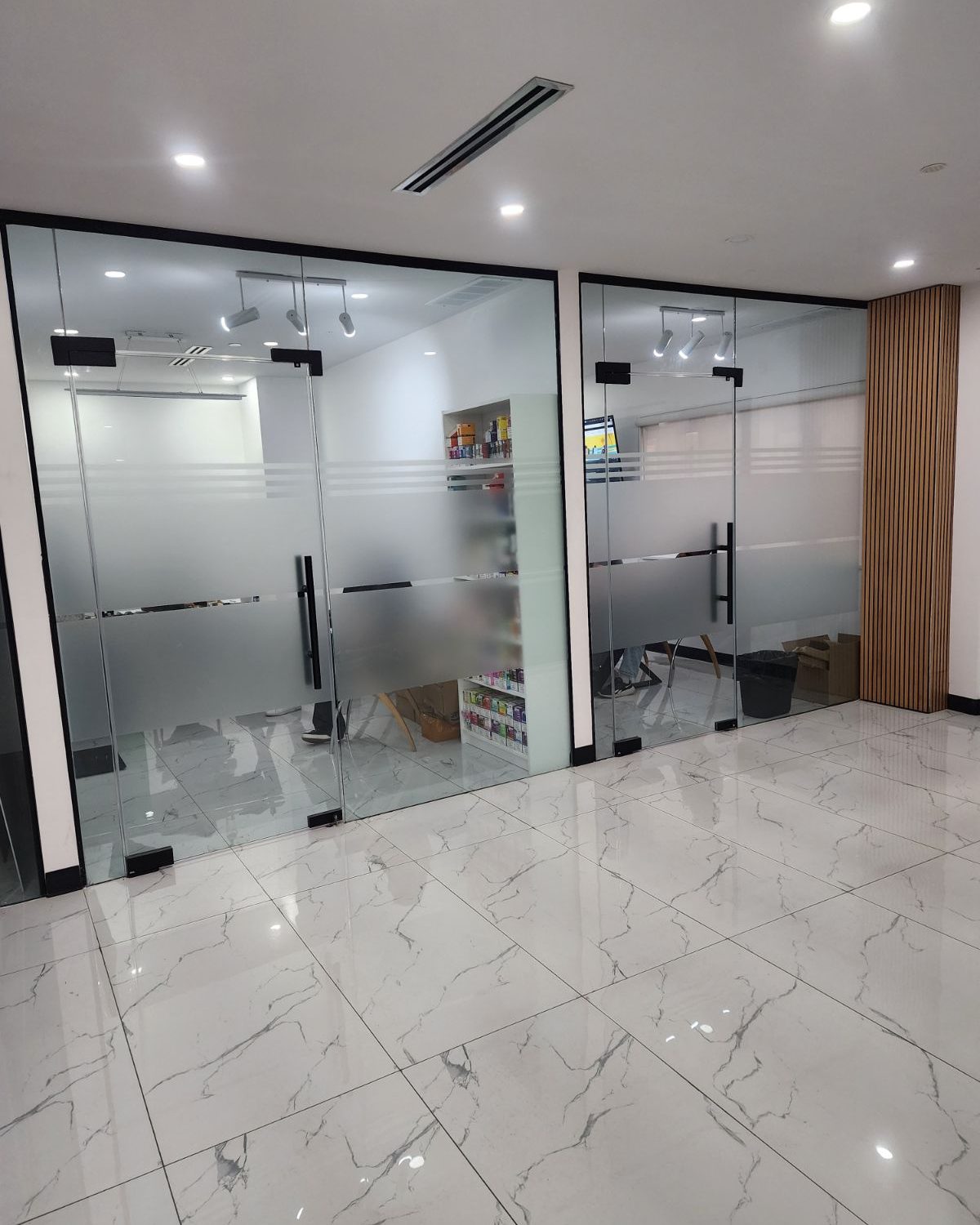 Two frameless glass door pairs on black patch fittings, set into frosted glass office fronts across a marble-floored corridor, installed by Canuck Door Systems