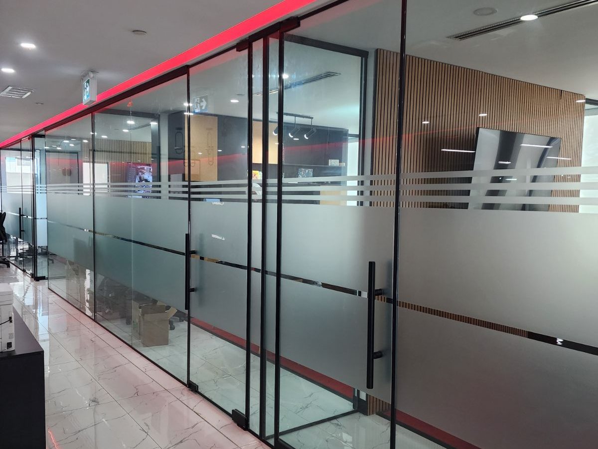 A glazed office front of frameless glass panels with frosted banding and a black ladder pull on the door leaf, running the length of an office corridor, installed by Canuck Door Systems