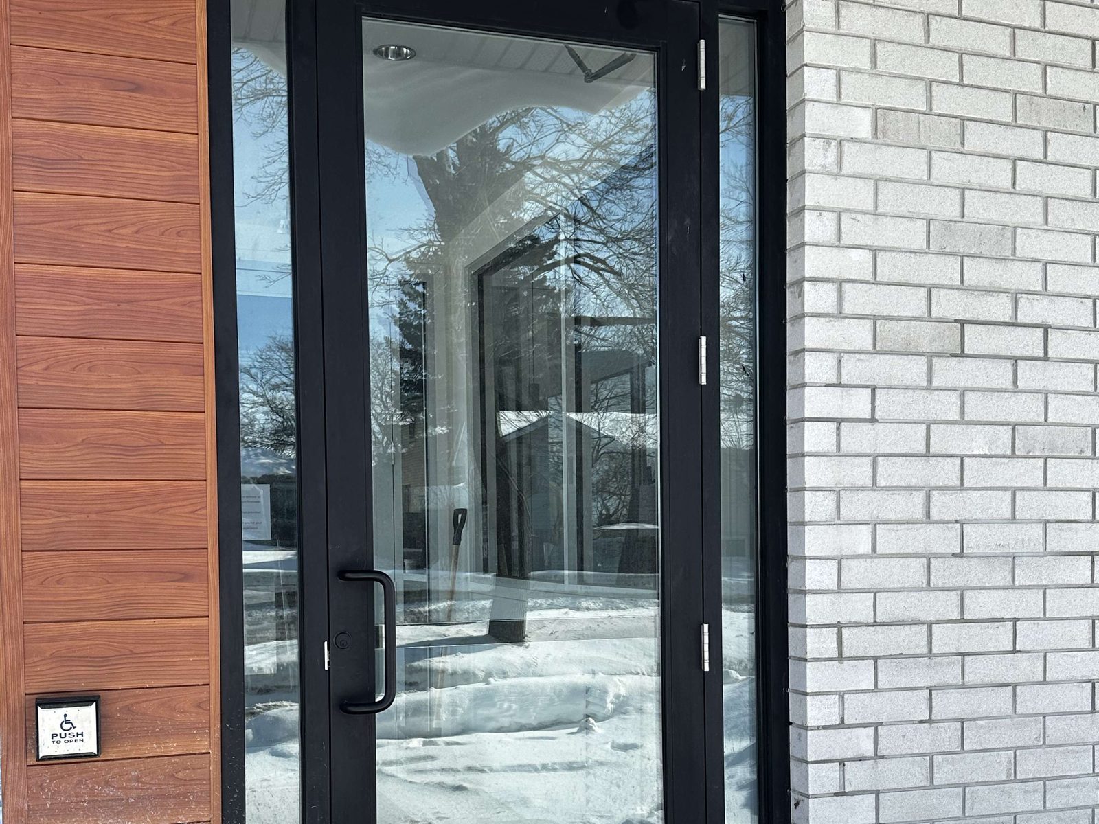 Black aluminum full-glass entrance door on a continuous hinge with a ladder pull, a sidelite beside it and a push-to-open operator plate on the wall, installed and serviced by Canuck Door Systems