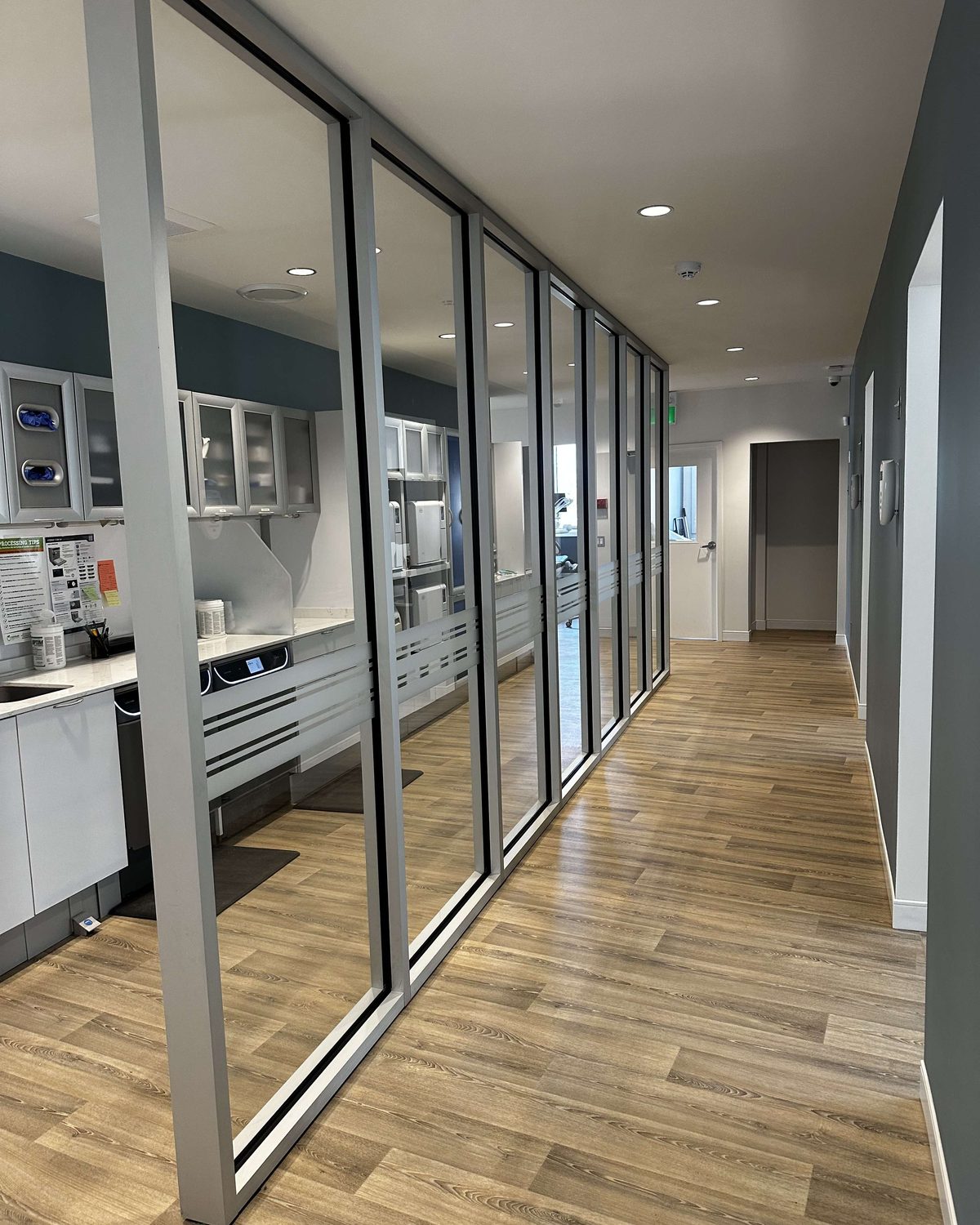 A glazed partition run in slim silver framing along a clinic corridor with a wood-look floor, the sterilization room visible through the glass, installed by Canuck Door Systems