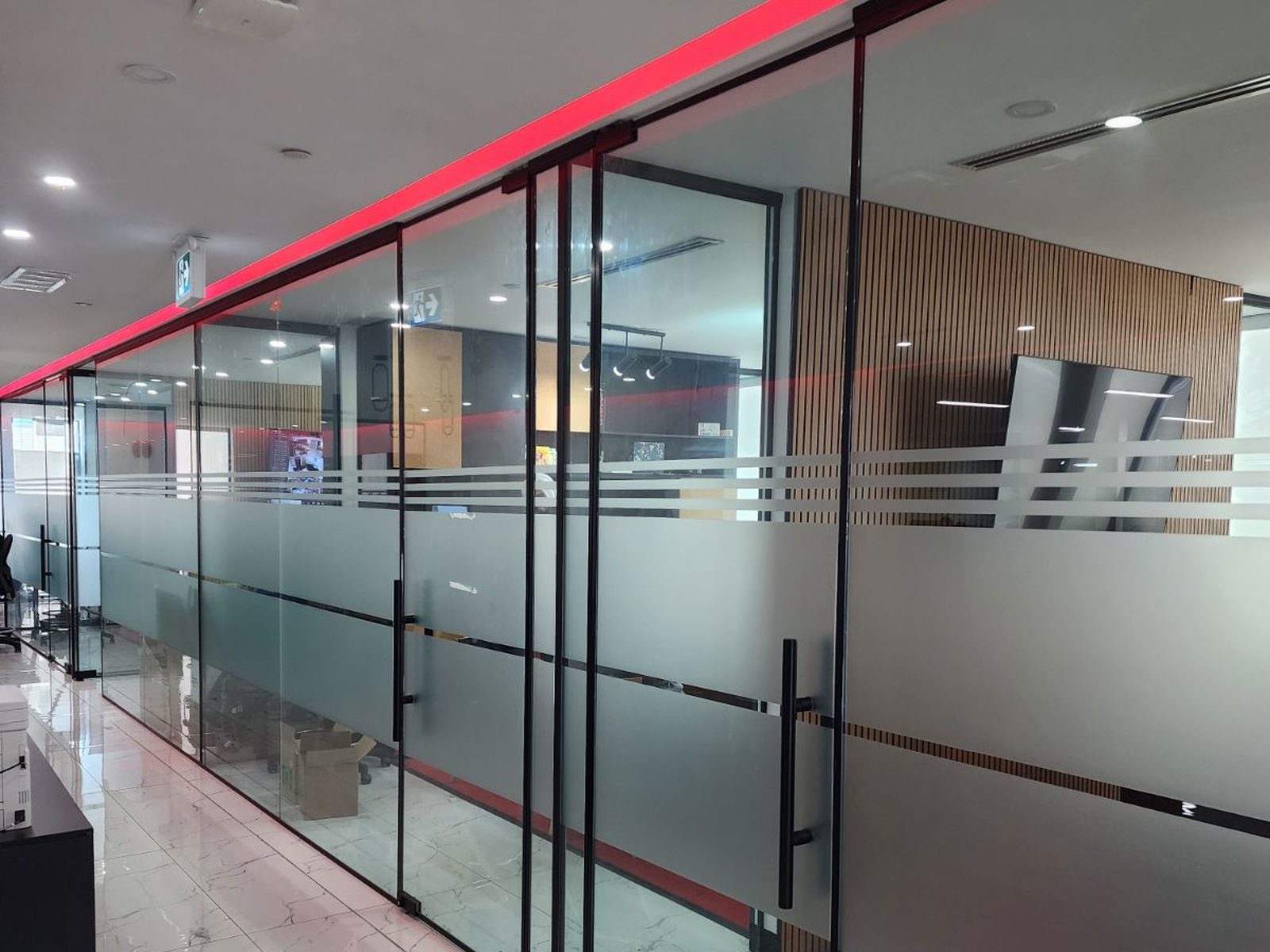 A long run of full-height glass partition walls in slim black framing along an office corridor, with a glass door on a black ladder pull and frosted manifestation banding across the glass, installed by Canuck Door Systems