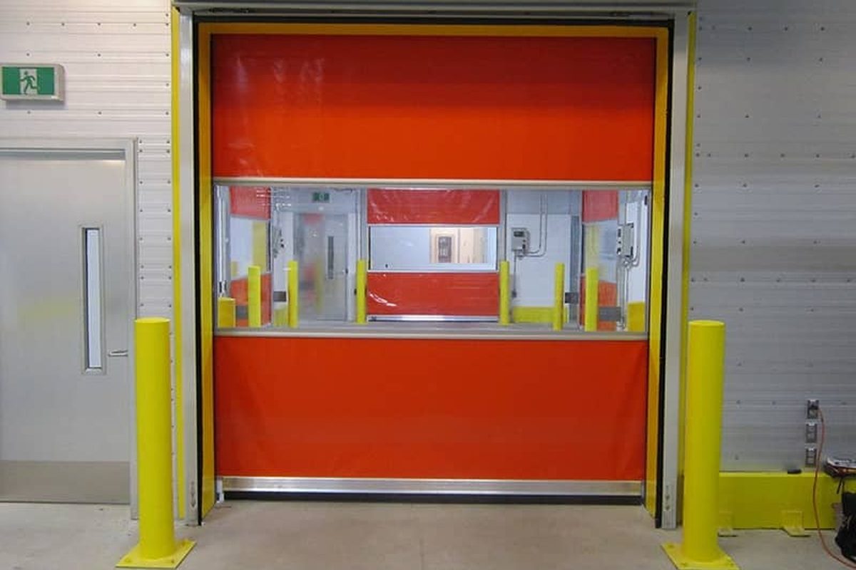 An orange high-speed fabric door closed in an interior industrial opening between yellow bollards, with a full-width vision panel across the curtain