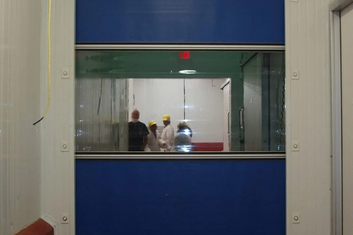 A blue high-speed fabric door closed across a plant opening, its vision window showing staff in hard hats working on the other side