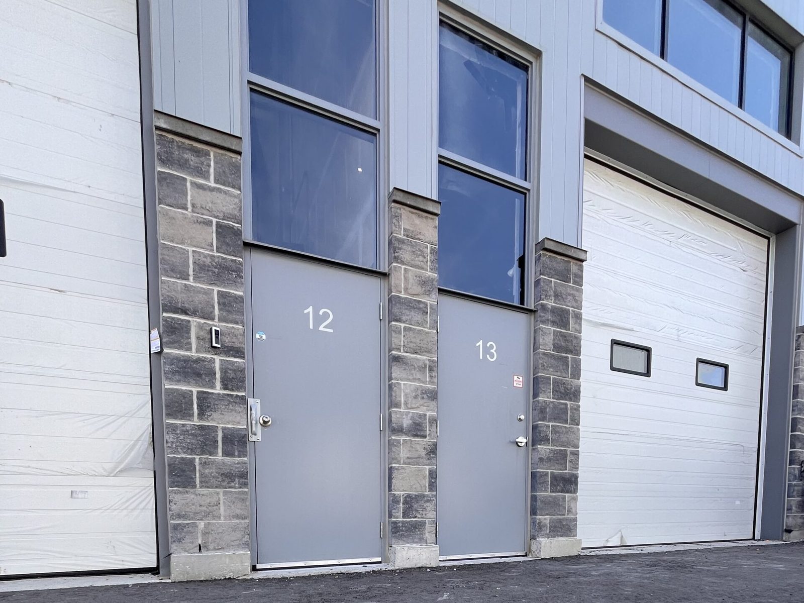 Two grey hollow metal personnel doors with glazed transoms, set in stone piers between insulated sectional overhead doors on an industrial unit, supplied and installed by Canuck Door Systems