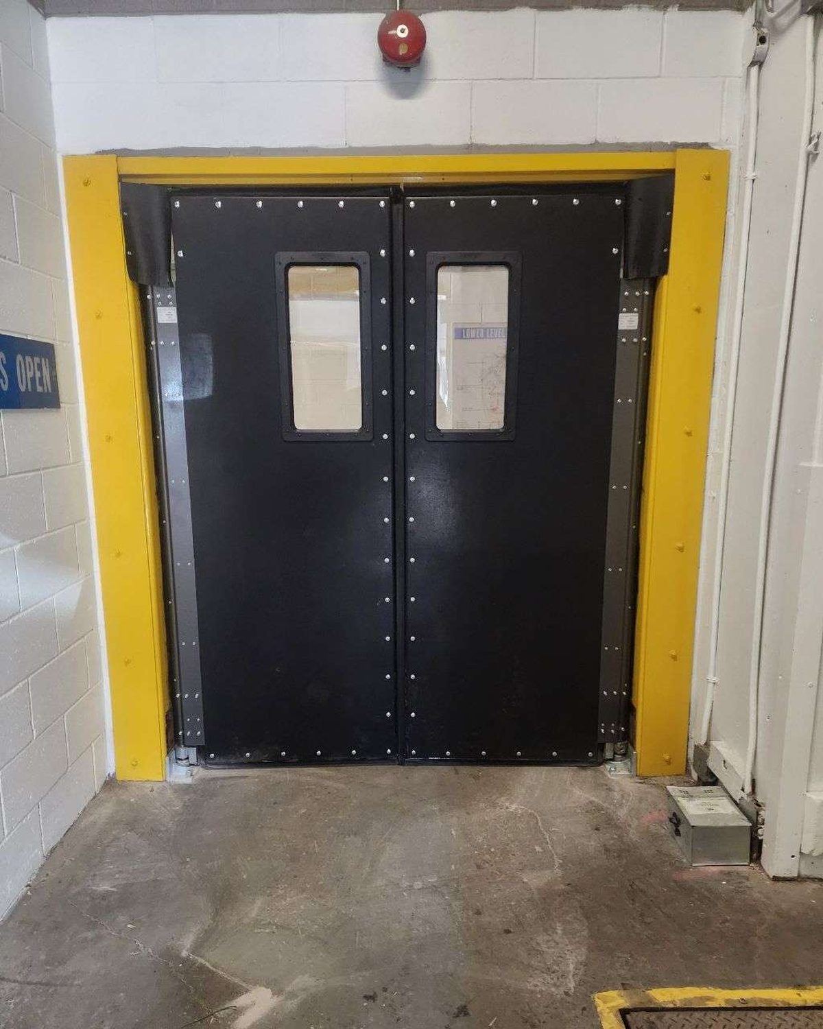 A pair of black double-acting impact traffic doors with vision panels, hung in a service corridor between yellow steel jamb guards, installed by Canuck Door Systems