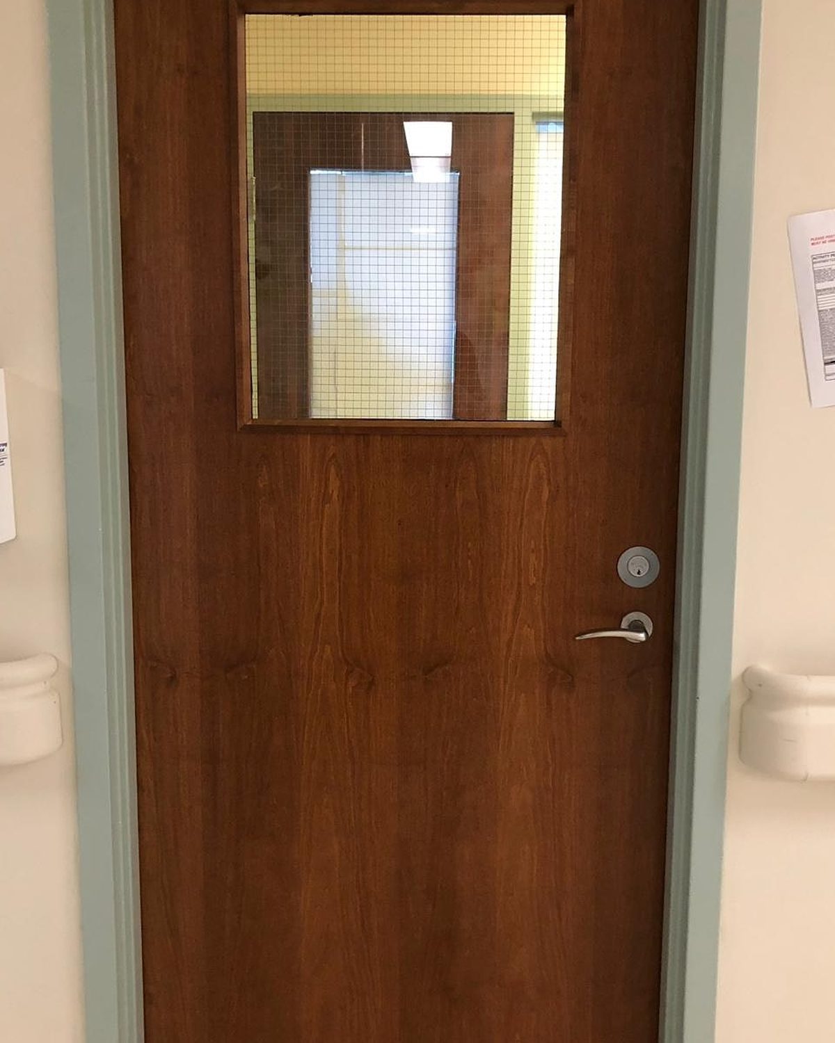A dark wood-veneer hospital door with a wired-glass vision panel and lever hardware in a patient-care corridor, installed by Canuck Door Systems