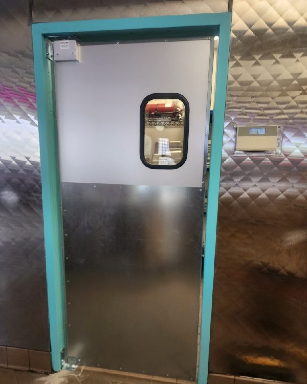 A commercial kitchen traffic door with a rounded vision window and brushed stainless lower panel, installed by Canuck Door Systems